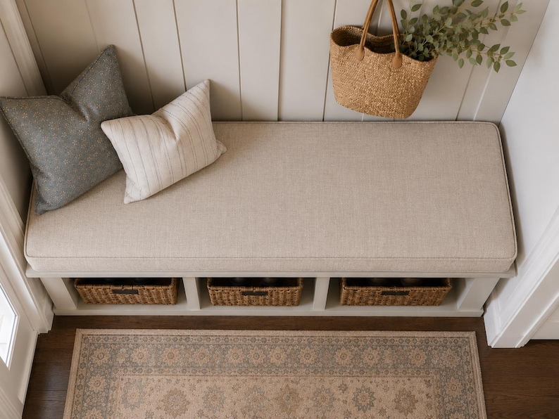 May include: A white entryway bench with a beige upholstered seat cushion, featuring three woven storage baskets underneath. Two decorative pillows, one solid gray and one white with thin stripes, sit on the bench alongside a hanging straw tote bag.