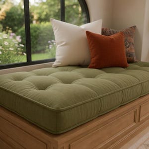 May include: A rectangular olive green tufted bench cushion with a textured fabric finish. The cushion is placed on a wooden base and features one large white square pillow and one smaller rust-colored square pillow resting on top of it.