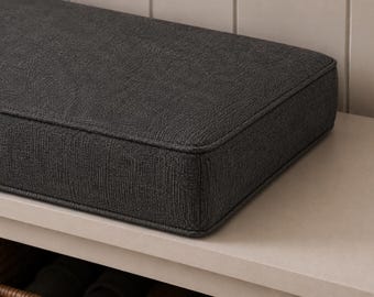 May include: A rectangular bench cushion upholstered in a dark charcoal gray fabric with a visible woven texture. The cushion features clean, structured edges with piped seams along the top and bottom perimeter, creating a firm and tailored appearance.