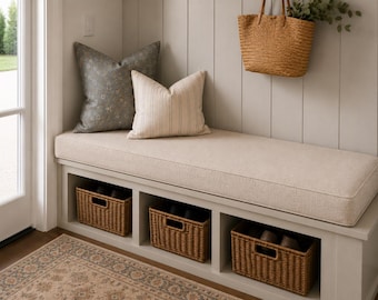 May include: A light beige upholstered bench with three open storage compartments containing woven brown wicker baskets. The bench features a gray floral patterned pillow and a cream striped pillow, with a woven straw tote bag hanging above.