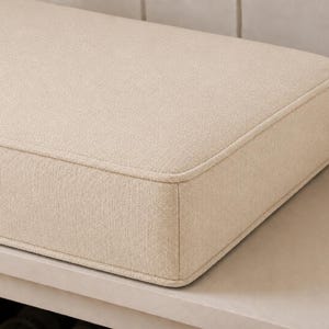 May include: A rectangular beige cushion with a textured fabric finish and piped edges. The cushion features a structured, boxy shape with rounded corners, showcasing a consistent woven pattern across its surface and sides, providing a clean look.