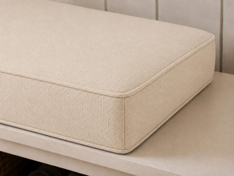 May include: A rectangular beige cushion with a textured fabric finish and piped edges. The cushion features a structured, boxy shape with rounded corners, showcasing a consistent woven pattern across its surface and sides, providing a clean look.