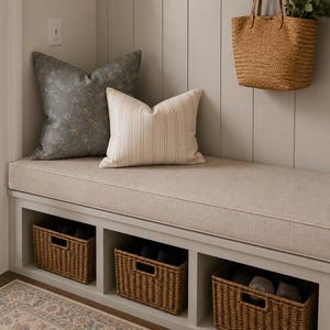 May include: A light beige upholstered bench with three open storage compartments containing woven wicker baskets. The bench features a large dark gray floral patterned pillow and a smaller cream-colored striped pillow, with a straw tote bag hanging above.