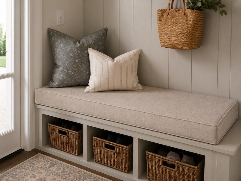 May include: A light beige upholstered bench with three open storage compartments containing woven wicker baskets. The bench features a large dark gray floral patterned pillow and a smaller cream-colored striped pillow, with a straw tote bag hanging above.