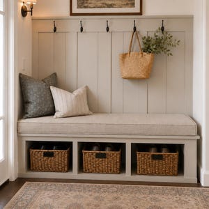 May include: A light gray entryway bench featuring a vertical paneled backrest with five black metal hooks, a beige upholstered seat cushion, and three open storage cubbies containing woven wicker baskets with black metal label holders.