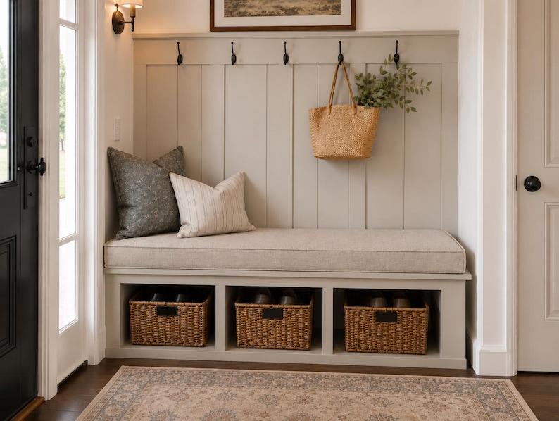 May include: A light gray entryway bench featuring a vertical paneled backrest with five black metal hooks, a beige upholstered seat cushion, and three open storage cubbies containing woven wicker baskets with black metal label holders.