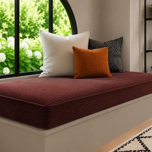 May include: A rectangular window seat cushion upholstered in a deep burgundy corduroy fabric. The cushion is topped with three decorative throw pillows in solid white, solid burnt orange, and a black and white geometric patterned fabric.