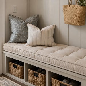 May include: A light beige tufted bench cushion sits atop a white storage bench with three open cubbies containing woven seagrass baskets. Two decorative throw pillows, one dark gray floral and one beige striped, rest on the bench seat.
