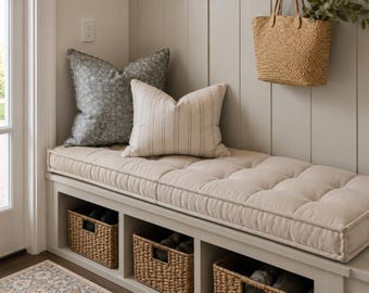 May include: A light beige tufted bench cushion sits atop a white storage bench with three open cubbies containing woven seagrass baskets. Two decorative throw pillows, one dark gray floral and one beige striped, rest on the bench seat.
