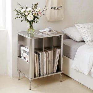 May include: A stainless steel bedside table with a flat top surface and three vertical storage compartments. The unit features a side pocket holding a magazine labeled FRAME and interior shelves filled with various books and magazines.