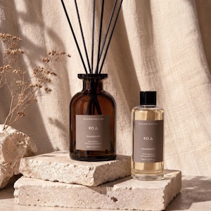Puede incluir: Un frasco difusor de varillas de vidrio marrón con varillas negras y una botella de recambio de vidrio transparente a juego, ambos con etiquetas que dicen Skandinavisk RO Tranquility 200ml/6.76 fl. oz. Los frascos están sobre bloques de piedra.