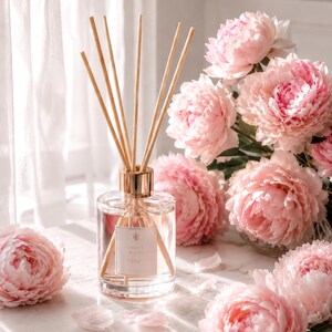 May include: A clear glass reed diffuser bottle with a gold-toned collar and several wooden reeds inserted. The label on the front of the bottle features the text "PEONY BLUSH" in a simple font. The diffuser is surrounded by several large, light pink peony flowers.