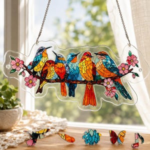 Puede incluir: Un atrapasol colgante con una ilustración estilo mosaico de cinco pájaros coloridos posados en una rama florida. Los pájaros muestran patrones vibrantes en azul, naranja, amarillo y rojo. Flores rosas acentúan la rama. Incluye piezas sueltas.