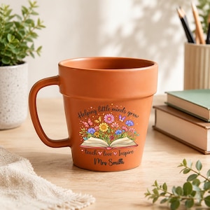 Può includere: Una tazza in ceramica color terracotta a forma di vaso da fiori con manico. La parte anteriore presenta un grafico di un libro aperto con fiori colorati e farfalle che emergono dalle pagine, con il testo "Helping little minds grow", "teach love Inspire" e "Mrs Smith".