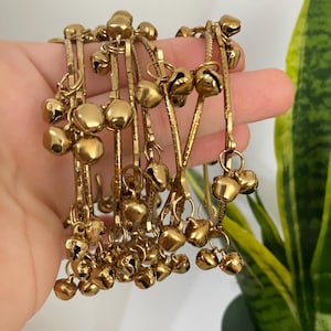 Könnte beinhalten: Ein Set aus goldfarbenen Metallarmreifen mit mehreren kleinen, runden Glöckchen, die an den dünnen, strukturierten Reifen befestigt sind. Die metallischen Armbänder sind in einem Bündel angeordnet und zeigen die glänzende, reflektierende Oberfläche der Glöckchen sowie das strukturierte, geriffelte Design der kreisförmigen Metallrahmen.