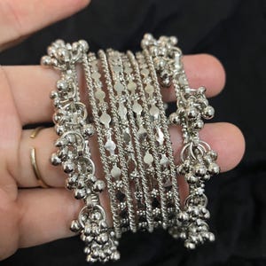 May include: A set of silver-toned metal bangles featuring a stack of five bracelets. The two outer bangles are adorned with small, dangling spherical charms, while the three inner bangles display a textured, repeating geometric pattern.