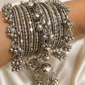 May include: A set of silver-toned metal bangles featuring a mix of textured, twisted, and smooth bands. The stack includes multiple dangling charms shaped like small bells and larger jhumka-style pendants that hang from the circular bracelets.
