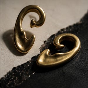 May include: A pair of gold-toned ear cuffs with a smooth, curved, and organic shape. Each piece features a polished metallic finish with a rounded, swirling design that tapers to a point, creating a continuous, fluid silhouette for the ear.