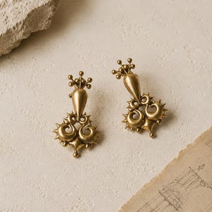 May include: A pair of gold-toned metal earrings featuring a teardrop-shaped top section topped with small spheres, connected to a lower section with swirling, spiked, and circular decorative elements in a symmetrical, ornate design.