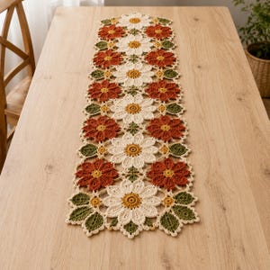 Könnte beinhalten: Ein rechteckiger Häkel-Tischläufer mit einem sich wiederholenden Blumenmuster. Das Design besteht aus abwechselnden cremefarbenen und rostfarbenen Gänseblümchenmotiven mit gelben Zentren, die durchgehend mit kleinen grünen Blattakzenten durchsetzt sind.