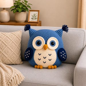 May include: A blue and cream crocheted owl pillow featuring large round eyes, a small orange beak, dark blue wings with white stitching, and two tufted ear accents. The owl has a cream-colored belly with dark blue chevron patterns and small orange feet.