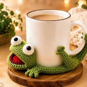 May include: A white ceramic mug held by a green crocheted frog cozy. The frog features large white eyes with black pupils, a wide open mouth with a red interior, and four small limbs that wrap around the mug to secure it in place.