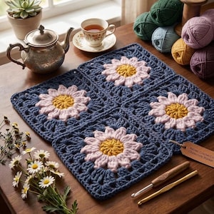 May include: A square crochet blanket composed of four joined granny squares, each featuring a central yellow and light pink daisy motif on a dark blue background. A brown leather tag reads Artisan Crochet Co. Chamomile Blanket, with two crochet hooks.