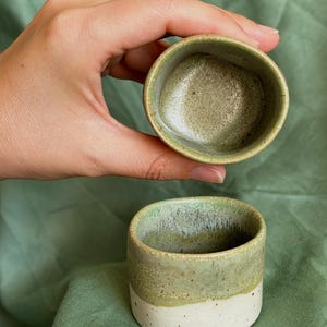 Può includere: Due piccole tazze in ceramica di forma cilindrica. La parte superiore di ogni tazza presenta uno smalto verde salvia maculato, mentre la parte inferiore è una base bianco sporco con puntini. L'interno è liscio e di colore verde.