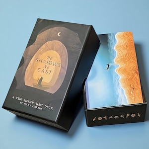 May include: A black rectangular tarot deck box titled The Shadows We Cast by Nicky Ferrada, featuring a silhouette illustration of a figure in a boat. Beside it, an open box reveals a card depicting a figure floating above a beach, labelled The Sun.