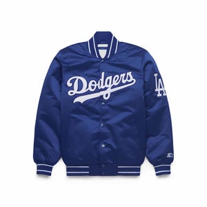 Puede incluir: Una chaqueta universitaria de satén azul real con el texto Dodgers en letra blanca en el pecho y un logotipo LA blanco en la manga izquierda. La chaqueta incluye un cierre frontal con botones a presión, cuello acanalado y puños a rayas azul y blanco.