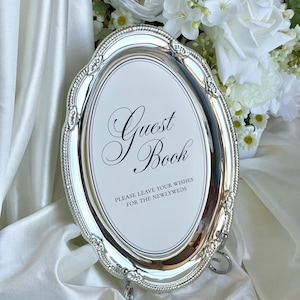 Może przedstawiać: Owalna ramka w kolorze srebrnym z ozdobnym, koralikowym brzegiem. Środkowa wkładka jest biała z czarnym napisem: Guest Book, PLEASE LEAVE YOUR WISHES FOR THE NEWLYWEDS. Ramka jest podparta małym, zintegrowanym metalowym stojakiem.