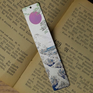 May include: A rectangular metal bookmark featuring a printed illustration of a mountain, stylized blue waves, green pine branches, and a large purple circle at the top. The bookmark has a small circular hole punched near the top edge for a ribbon.