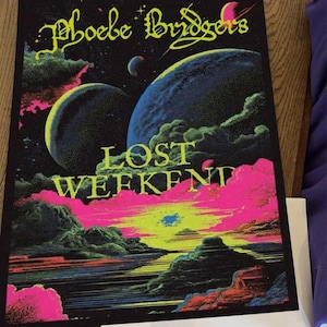May include: A rectangular poster featuring a space-themed illustration with the text Phoebe Bridgers and LOST WEEKEND in yellow. The artwork displays large blue planets, dark clouds, and a vibrant pink and yellow sunset over a dark landscape.