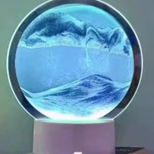 May include: A circular sand art lamp featuring a glowing blue light behind a glass disc filled with layered blue and white sand. The circular frame is mounted on a solid white cylindrical base with a small power button on the front surface.