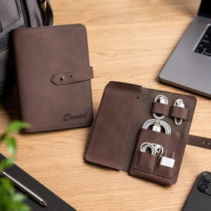 May include: A dark brown leather tech organizer case with a snap closure strap. One case is closed and features the name Daniel embossed on the front. The other case is open, revealing internal pockets holding white charging cables and a wall plug.