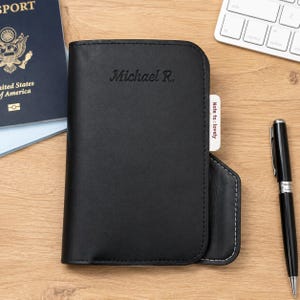 May include: A black leather passport holder featuring the embossed name Michael R. on the front cover. A small white tab extending from the side displays the printed text Note to lovely. The holder has rounded corners and visible stitching.