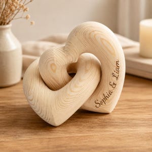 May include: A light-coloured wooden sculpture consisting of two interlocking heart shapes. The wood features a visible natural grain pattern. The text "Sophia & Liam" is engraved in a dark script font along the side of the larger heart shape.