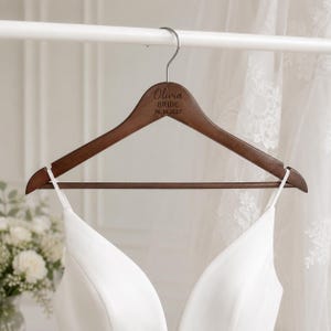 Puede incluir: Una percha de madera de color marrón oscuro con un gancho de metal plateado, que presenta el texto grabado Olivia BRIDE 06.14.2027. La percha incluye una barra horizontal y muescas, sosteniendo una prenda blanca con tirantes finos.