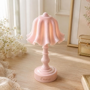 May include: A light pink table lamp featuring a ruffled, bell-shaped shade and a slender, tiered base. The circular base is engraved with the name Emma in a cursive script. The lamp has a smooth, glossy finish and a uniform pastel pink color.