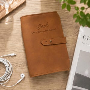 Peut inclure: Une couverture de carnet en cuir beige avec une sangle horizontale dotée d'une fermeture à bouton-pression en métal argenté. Le devant est gravé du nom Jack et du texte Stay organized, stay inspired. La couverture a une finition mate lisse.