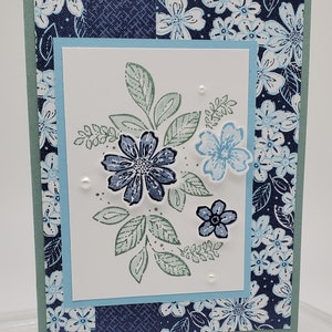 Op de afbeelding: Een kaart met een blauwe en witte bloemenprint op de rand. De kaart heeft een witte achtergrond met een blauw en groen bloemenontwerp in het midden. De tekst "Laura Borchert" en "www.LaurasStampPad.com" staat onderaan de kaart gedrukt.