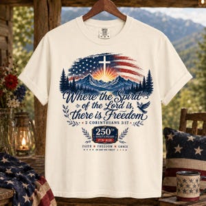 Könnte beinhalten: Ein cremefarbenes Kurzarm-T-Shirt mit einer Grafik aus amerikanischer Flagge, Bergen und einem Kreuz. Der Text lautet: Where the Spirit of the Lord is, there is Freedom, 2 Corinthians 3:17, 250th Anniversary 1776-2026, Faith, Freedom, Grace, In God We Trust.