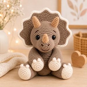 May include: A crocheted triceratops toy in shades of brown and beige. The dinosaur features a large frill with a white border, three light brown horns, dark round eyes, and white circular details on the feet. It is seated in an upright position.