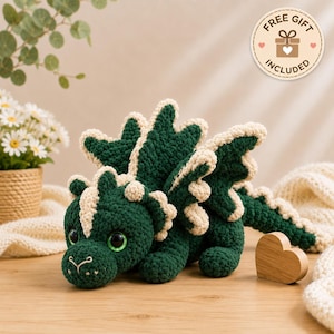 May include: A dark green and cream-colored crocheted dragon plush toy with large black eyes, textured scales along its back, and a long tail. A circular badge in the upper right corner features a gift icon and the text FREE GIFT INCLUDED.