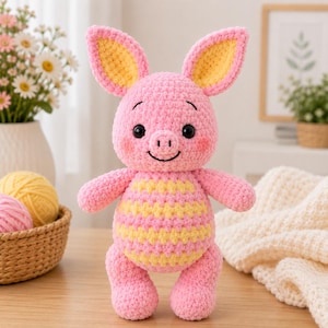 Könnte beinhalten: Ein gehäkeltes Stofftier in Form eines kleinen Schweinchens aus rosa Garn, mit großen aufrechten Ohren mit gelben Innenseiten und einem runden Körper mit horizontalen gelben und rosa Streifen. Das Spielzeug hat schwarze Kunststoffaugen und ein gesticktes Gesicht.