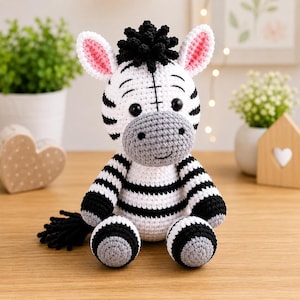 Può includere: Un peluche a forma di zebra realizzato all'uncinetto con corpo bianco, strisce nere, muso grigio e criniera nera. La zebra ha grandi occhi neri, interno delle orecchie rosa e una coda nera. Il giocattolo presenta punti di cucitura visibili.