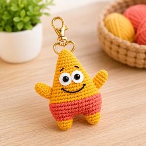 May include: A crochet keyring shaped like a star character with yellow skin and coral-coloured shorts. The figure features two black plastic eyes, embroidered eyebrows, and a stitched black smile. It is attached to a gold-toned metal lobster clasp.