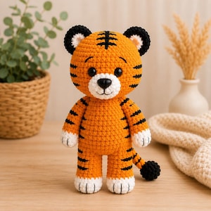 May include: A crochet tiger stuffed animal featuring bright orange yarn with black stripes, a white muzzle, and white paws. The toy has round black ears, black plastic eyes, and a black nose. It stands upright with a long tail ending in a black pom-pom.