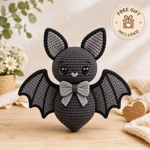 May include: A dark gray crocheted bat plush toy featuring large black safety eyes, a small light gray heart-shaped nose, and a light gray bow tie at the neck. The bat has pointed ears with light gray inner details and wide, spread wings.
