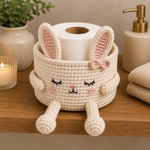 Puede incluir: Una cesta de ganchillo color crema con forma de conejo, orejas largas, un lazo pequeño y rasgos faciales bordados que incluyen ojos cerrados, nariz rosa y mejillas rosadas. La cesta contiene un rollo de papel higiénico y tiene dos piernas.